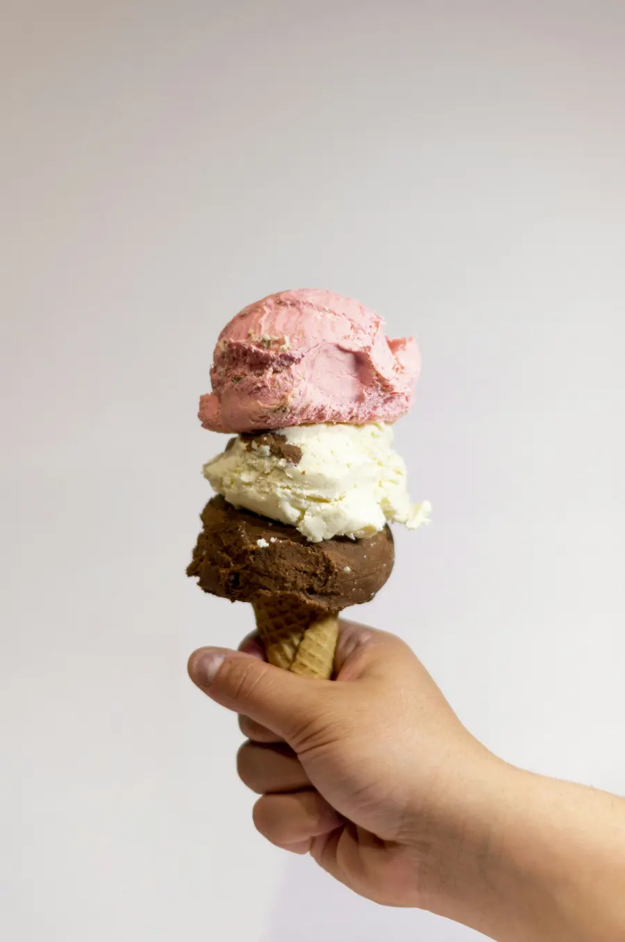 Mão segurando casquinha com três bolas: morango, creme e chocolate