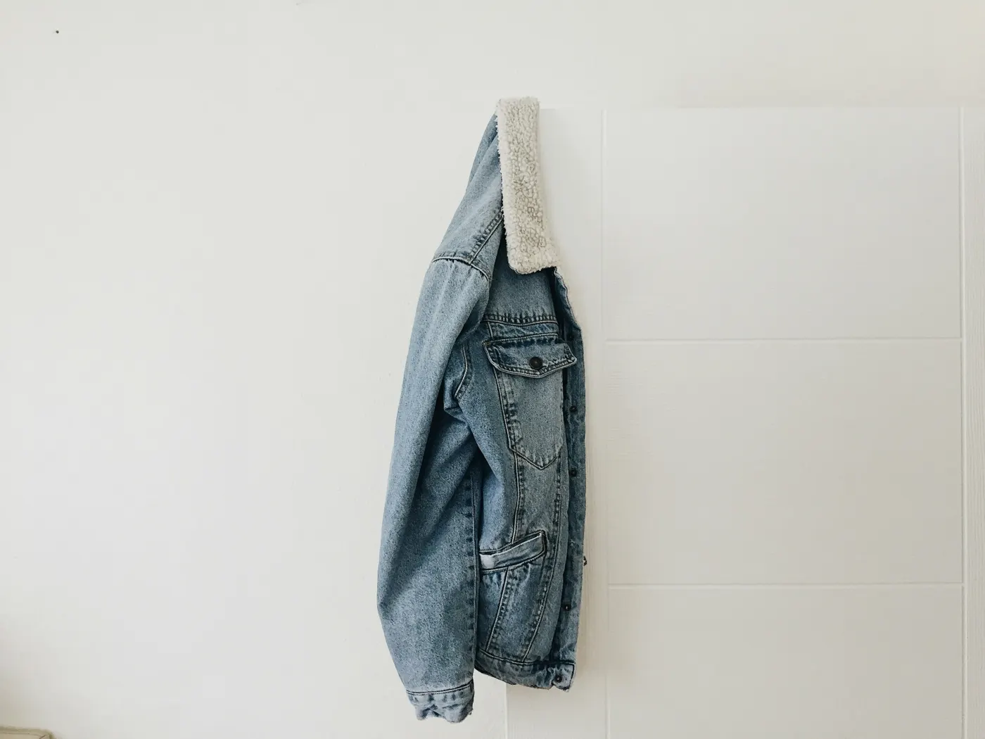 Jaqueta jeans clara com gola de pelúcia branca, pendurada em uma parede branca