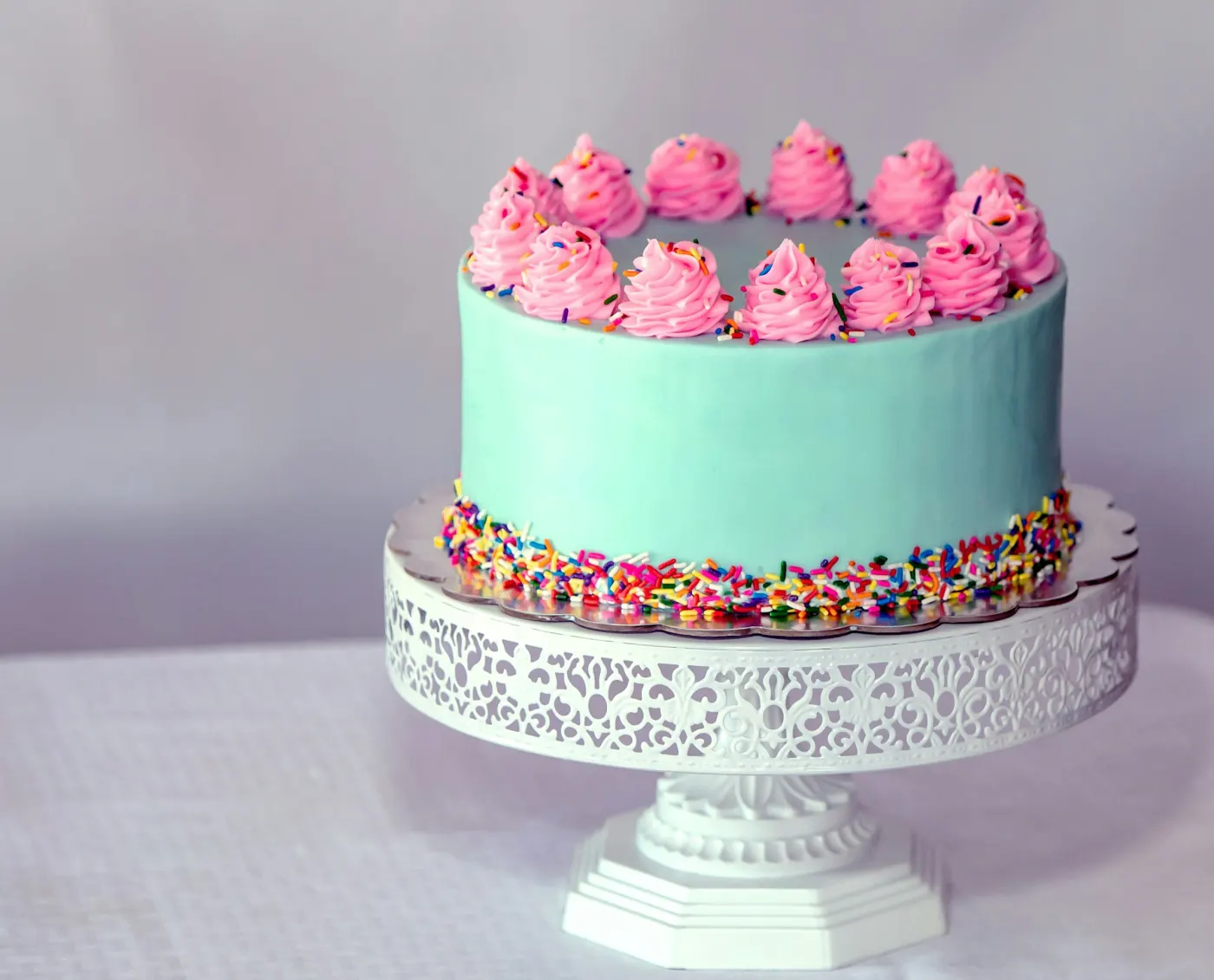 Bolo redondo de cobertura verde-água com bicos de chantilly rosa e granulado colorido, sobre um pedestal branco