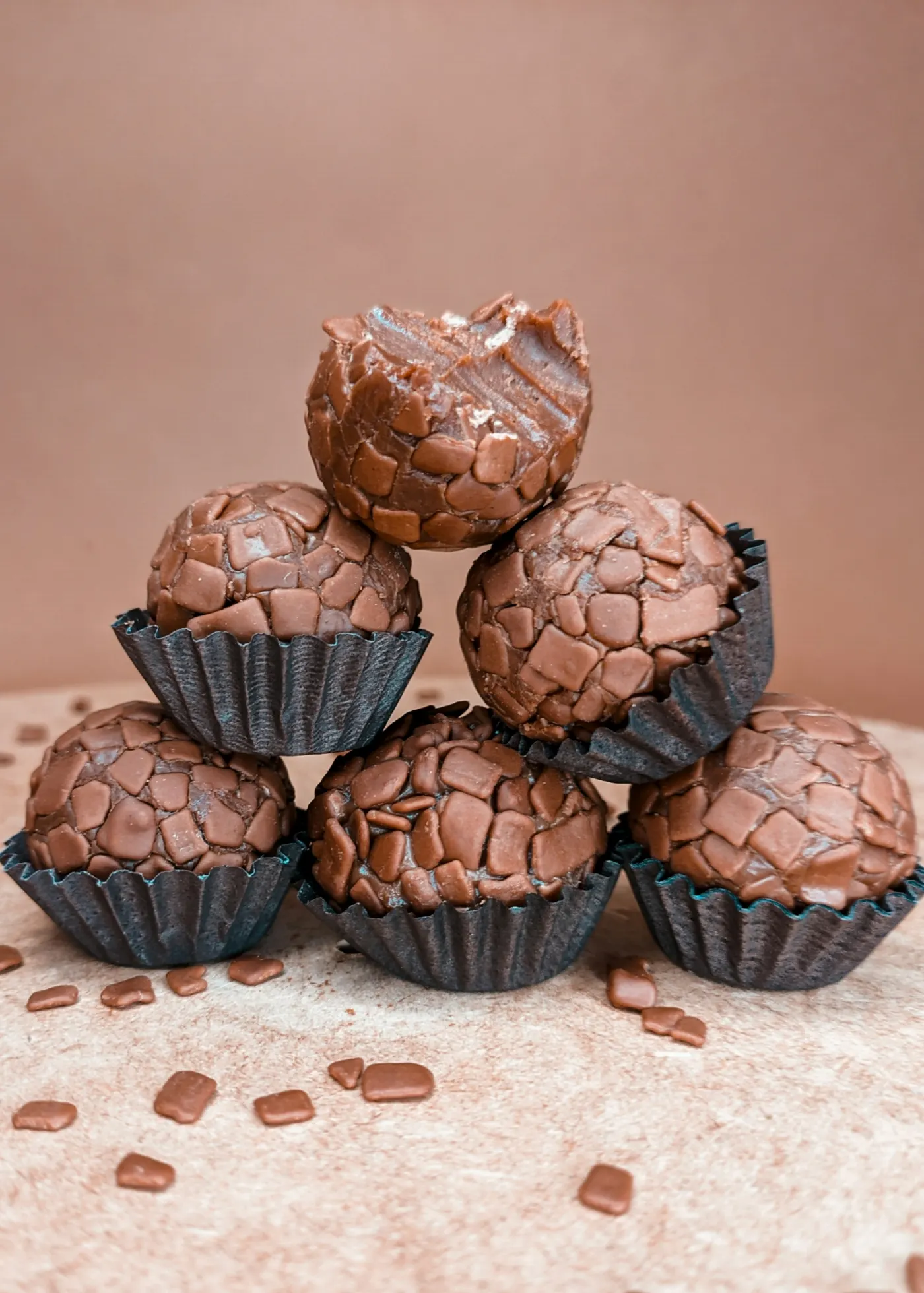 Seis brigadeiros gourmet cobertos de chocolate em lascas, empilhados em forminhas pretas, um deles com uma mordida
