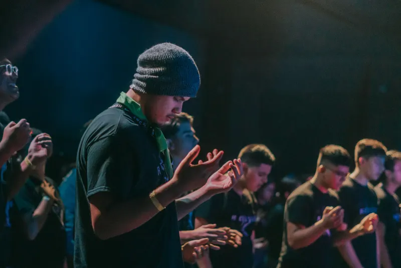 Homem de gorro, de mãos abertas, entre jovens num culto