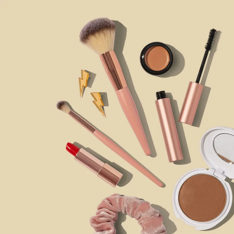 Pincéis rosa, batom, máscara de cílios, blush e um prendedor de cabelo sobre fundo pêssego