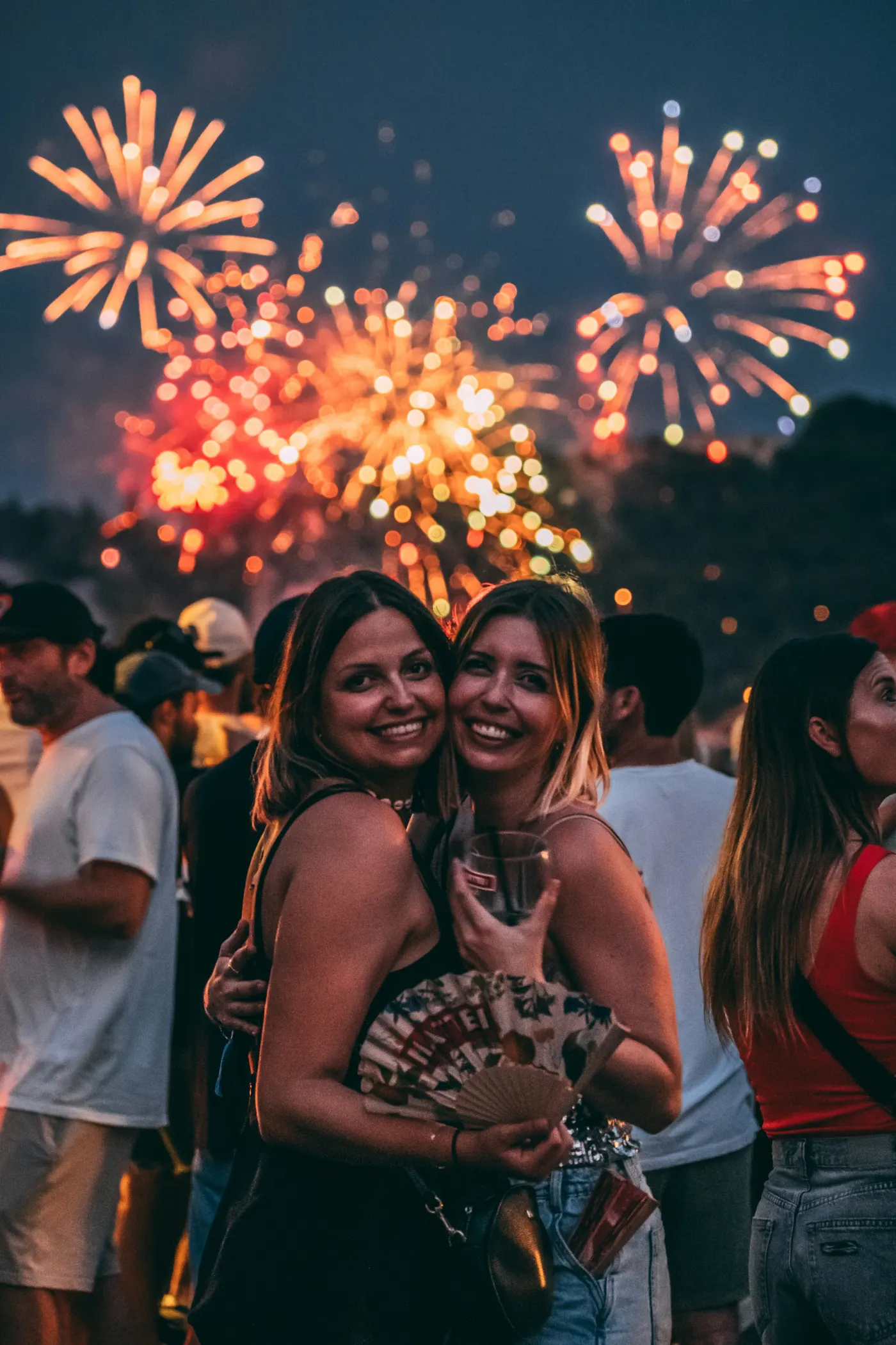 Duas amigas abraçadas e sorrindo, com fogos de artifício coloridos ao fundo