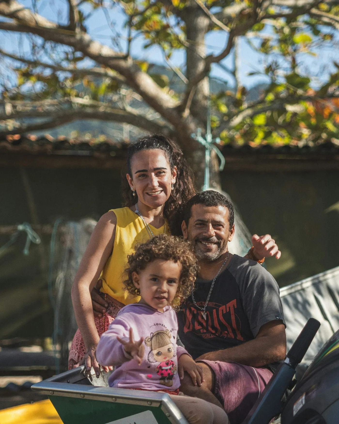 Pai, mãe e filha pequena sorrindo juntos ao ar livre, sob uma árvore
