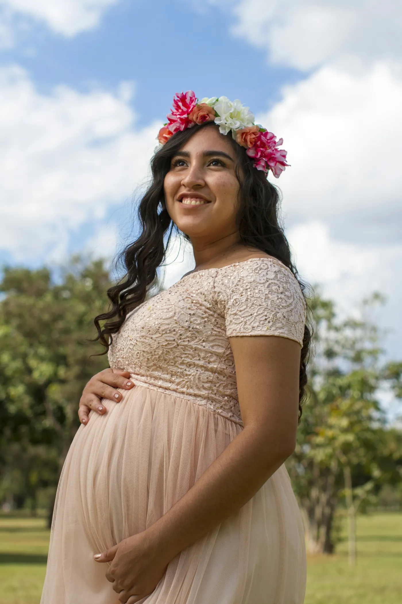 Gestante de coroa de flores e vestido rosa-claro, com as mãos na barriga, sorrindo para o céu