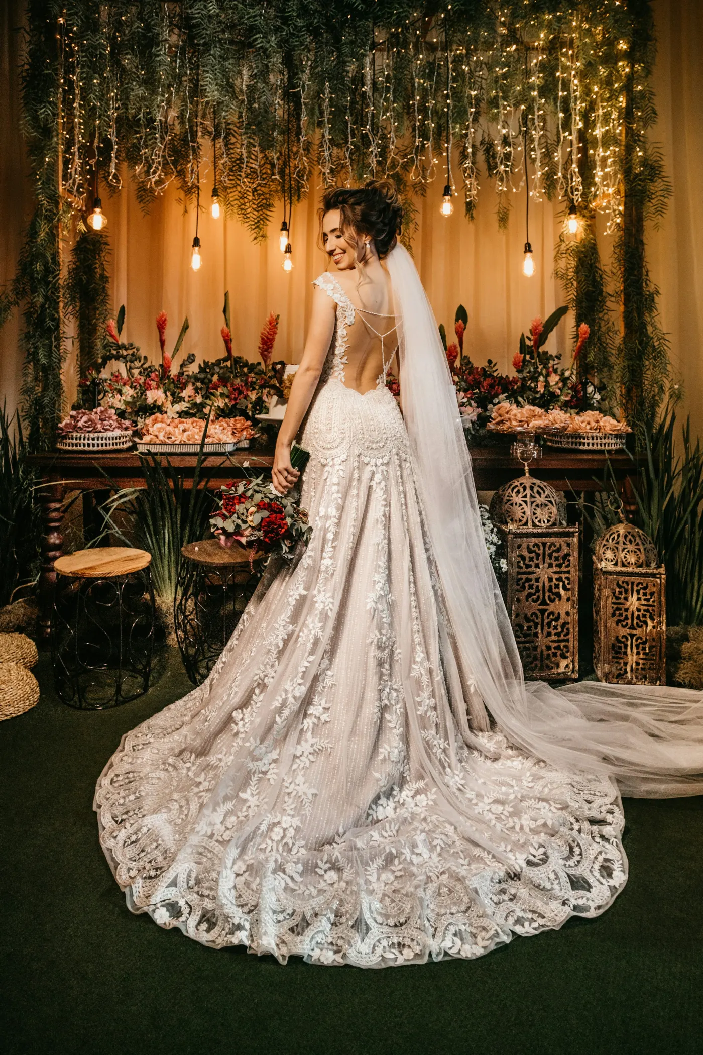 Noiva de costas com véu longo e vestido de renda, sorrindo para o lado diante de uma mesa de flores