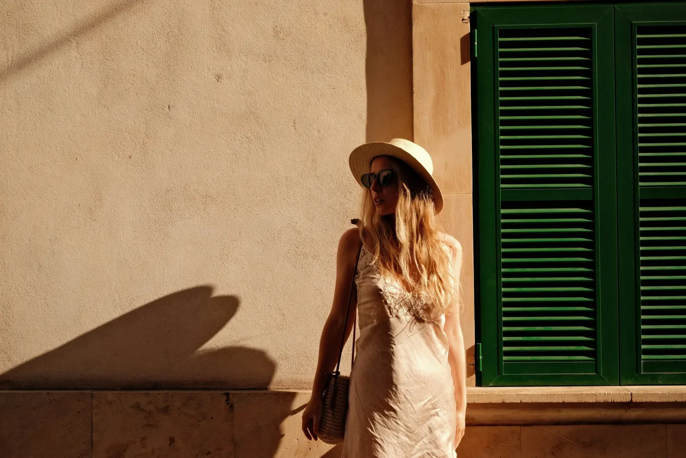 Mulher de chapéu de palha e vestido claro, encostada numa parede de sol com janela verde