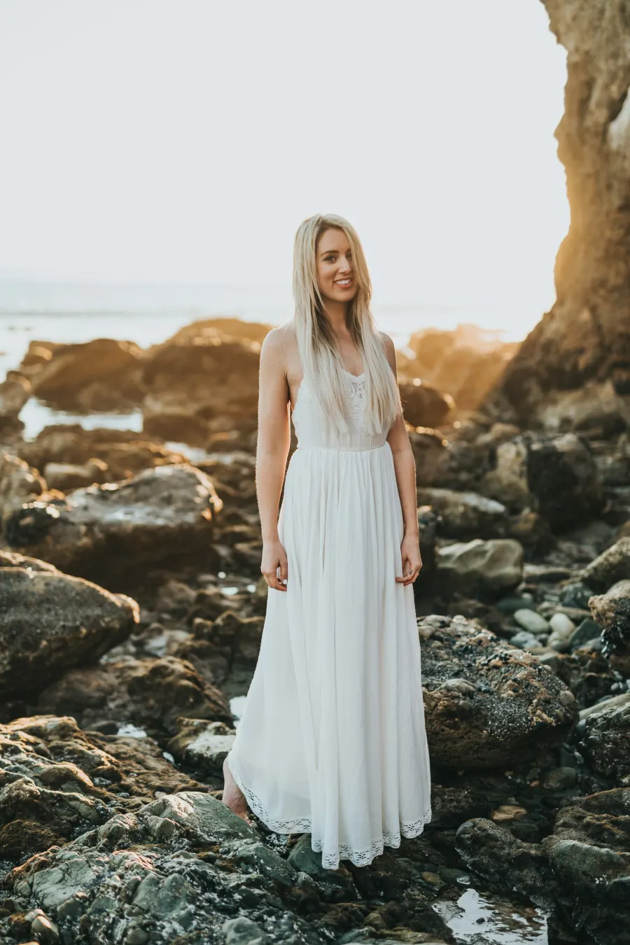 Mulher de vestido longo branco de alça fina, com renda na barra, sobre pedras à beira-mar