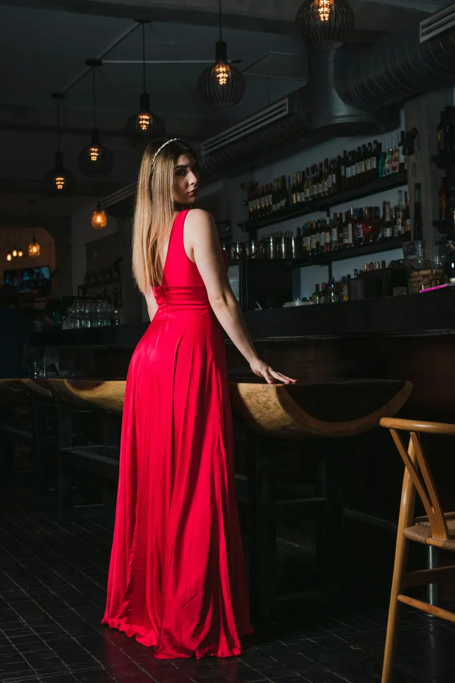 Mulher de costas, olhando por cima do ombro, com vestido longo vermelho de alça larga junto a um balcão de bar