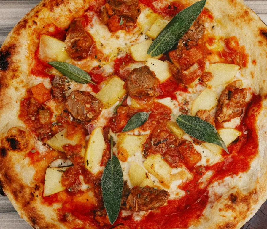 Pizza de borda manchada de lenha com carne de sol, macaxeira em cubos e folhas de sálvia, vista de cima