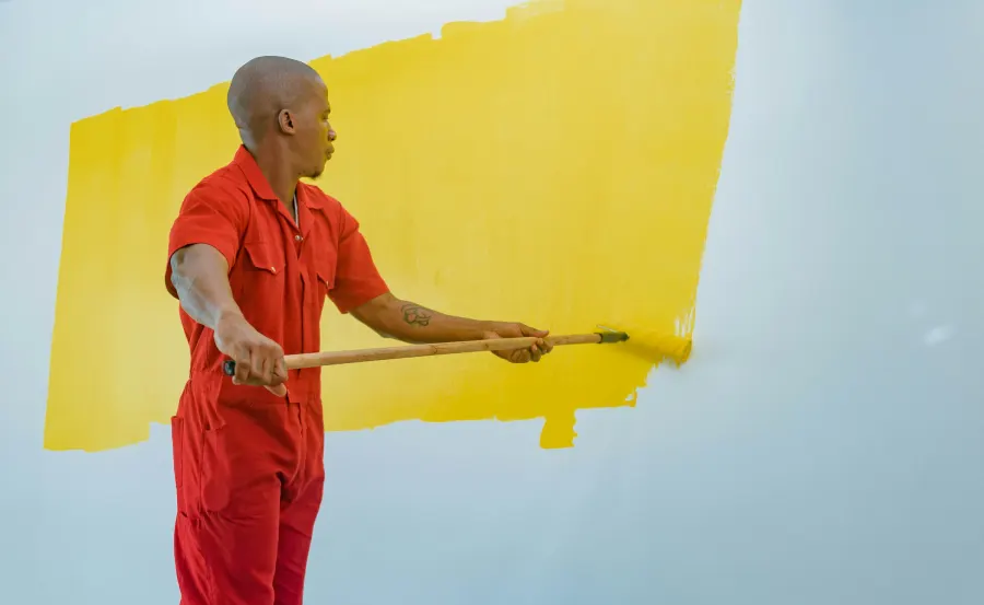 Pintor de macacão vermelho passando tinta amarela em uma parede clara com rolo de cabo longo