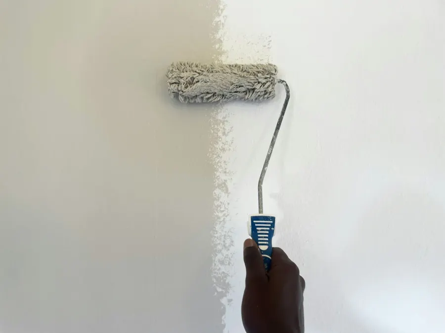 Mão passando rolo de tinta clara em uma parede, com metade já pintada