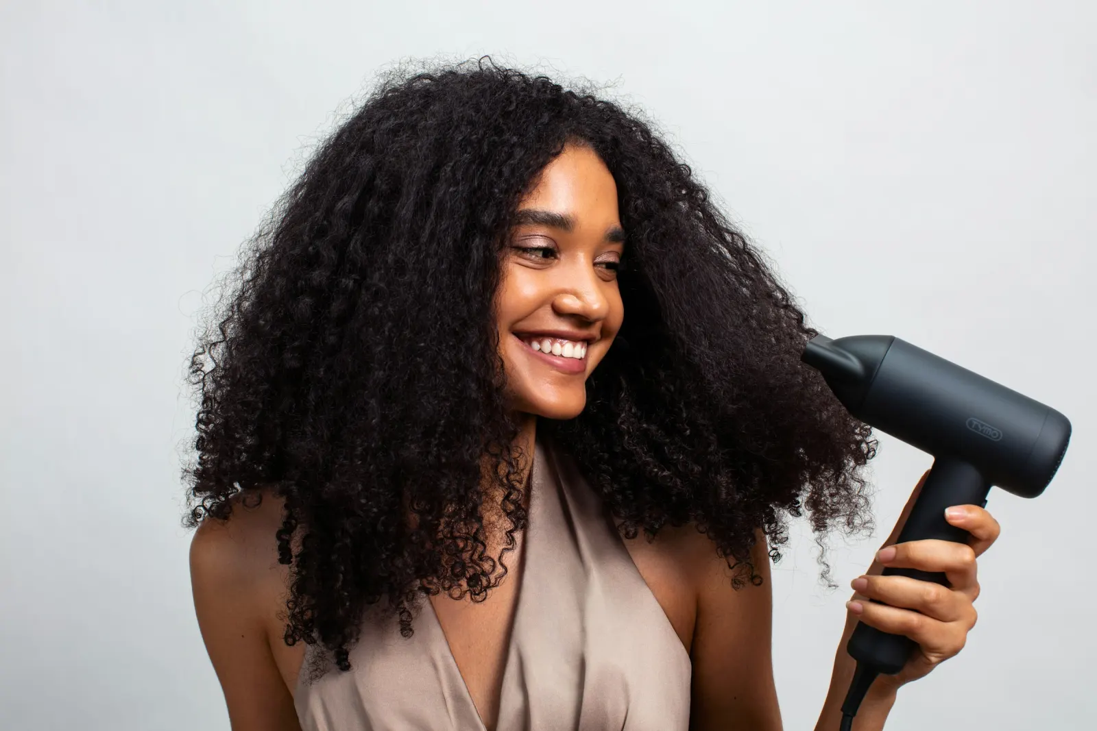 Mulher de cabelo cacheado volumoso sorrindo enquanto passa o secador