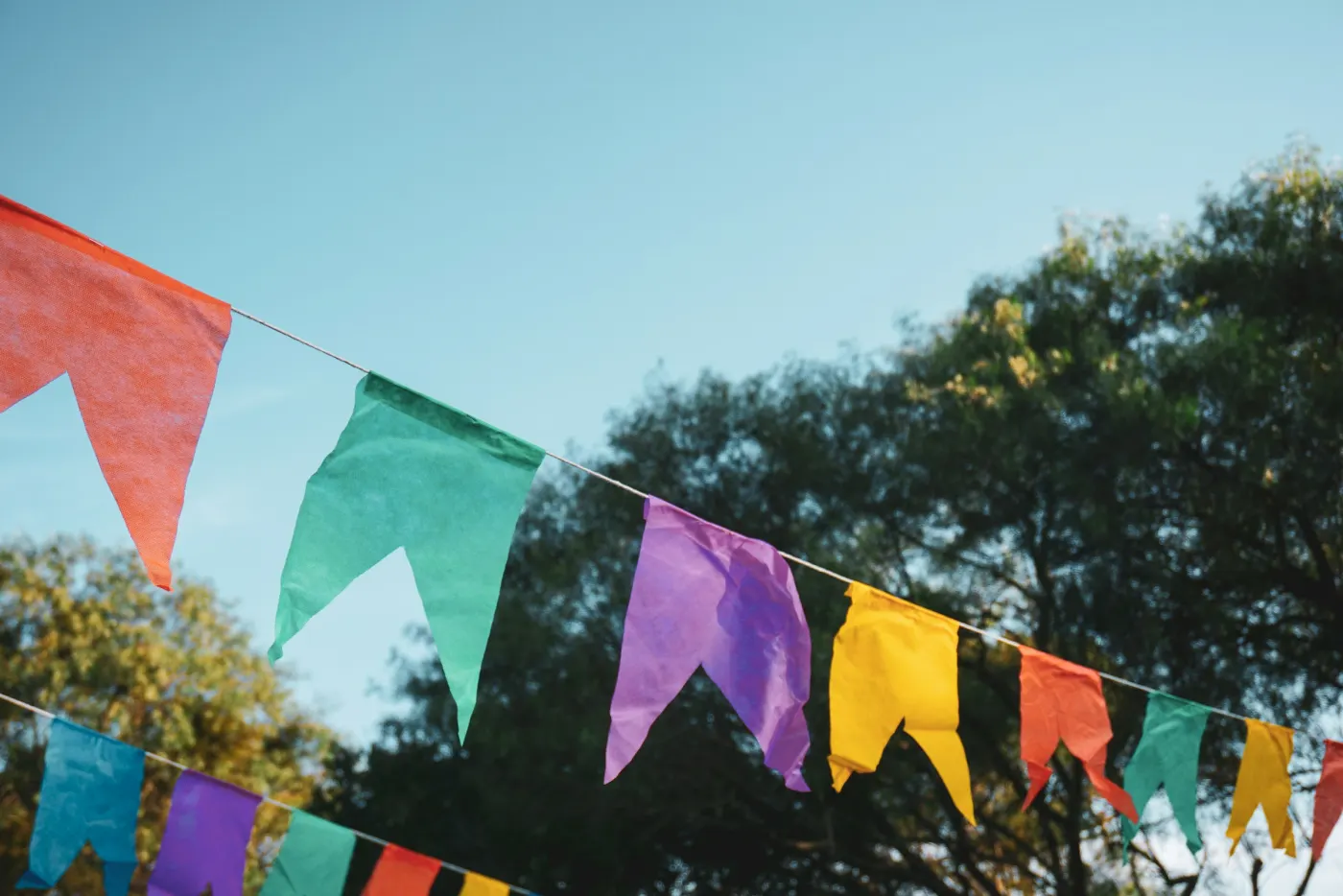 Cordões de bandeirinhas coloridas de papel, com árvores e céu azul ao fundo