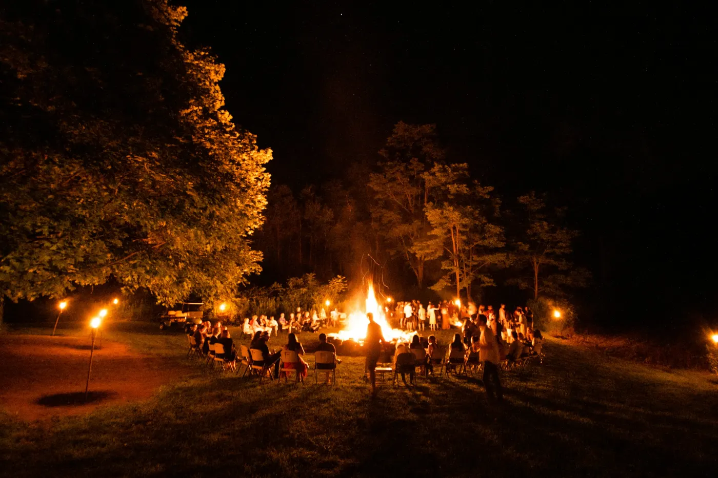 Fogueira acesa num terreno gramado à noite, com gente sentada em cadeiras em volta e tochas acesas