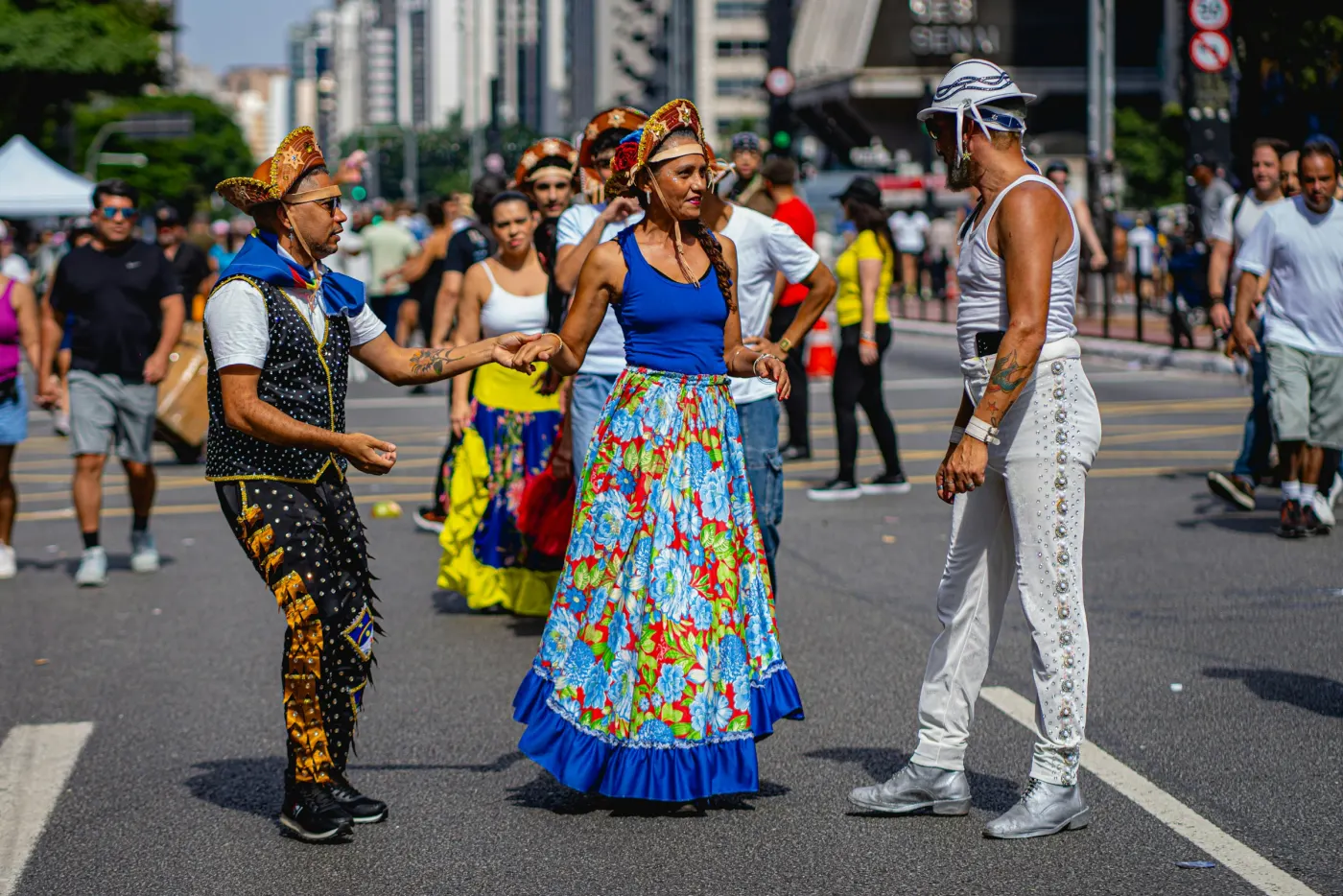 Dançarinos de quadrilha de chapéu de couro e saia florida, de mãos dadas numa rua cheia de gente