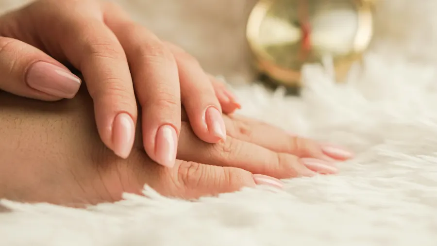 Duas mãos sobrepostas em um tapete de pelo branco, com unhas em gel de um rosa nude brilhante