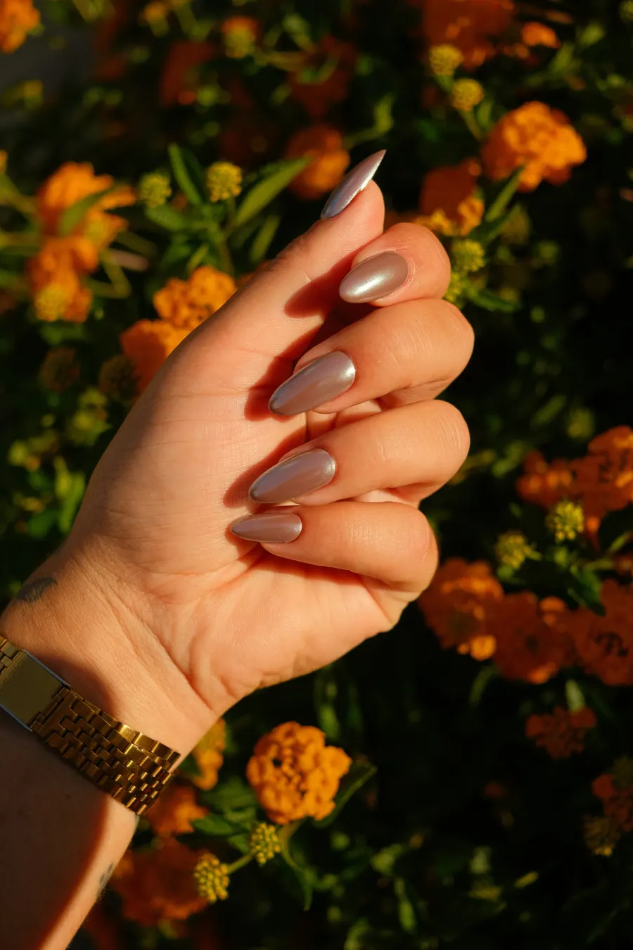 Mão com unhas amendoadas em nude perolado, diante de flores alaranjadas