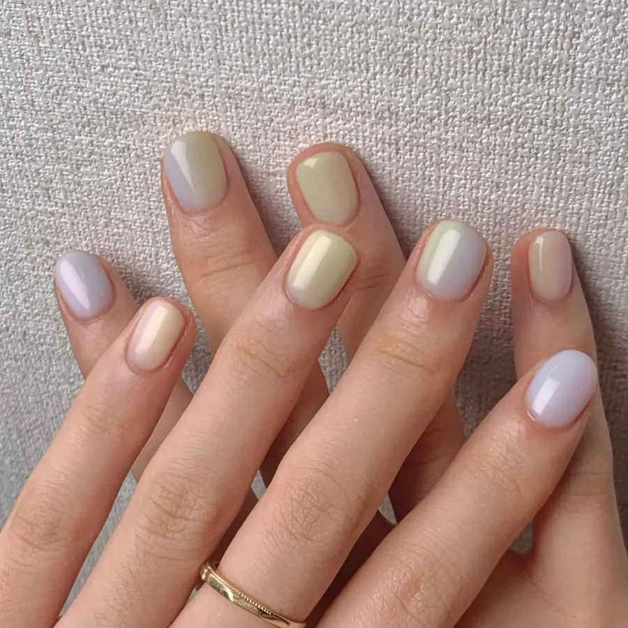 Duas mãos com unhas curtas em tons de nude e verde claro, e um anel dourado